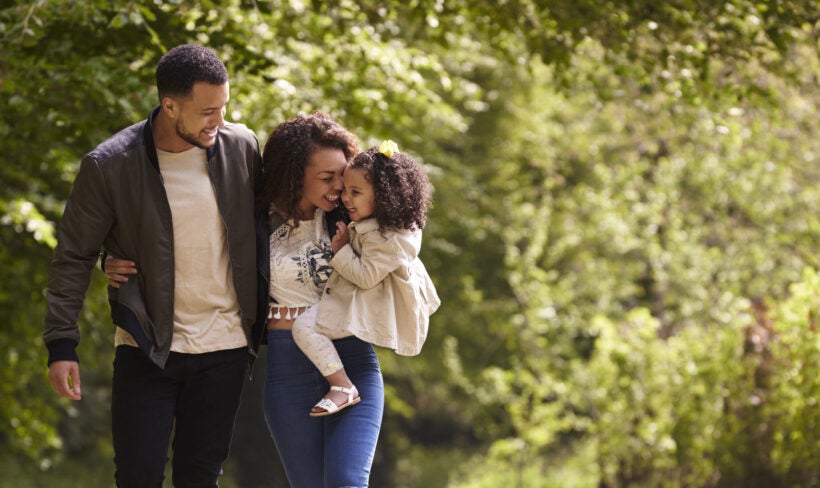 A smiling family in an outdoor setting with the mother holding her toddler and the father's arm is around the mother